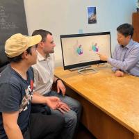 		The three researchers are sitting around a desk and Ailong Ke is pointing to an image of the IscB molecule on the computer screen.
	