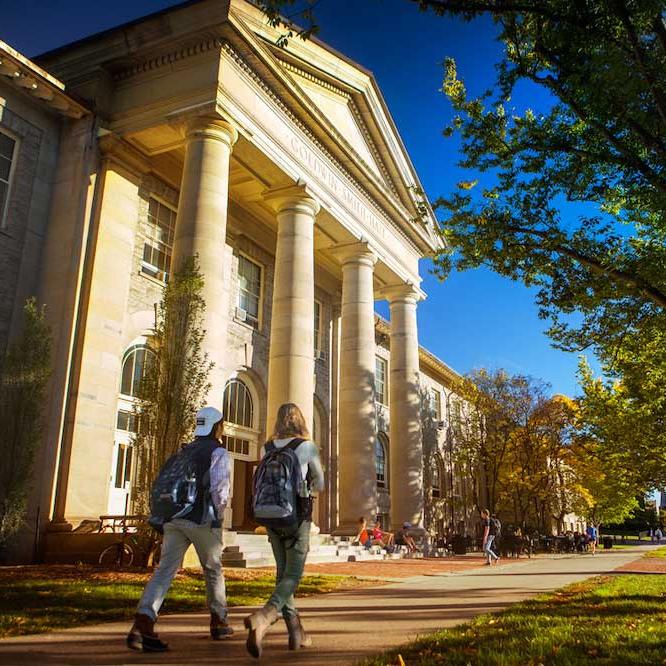 		Students with backpacks walk past a building with stone columns
	