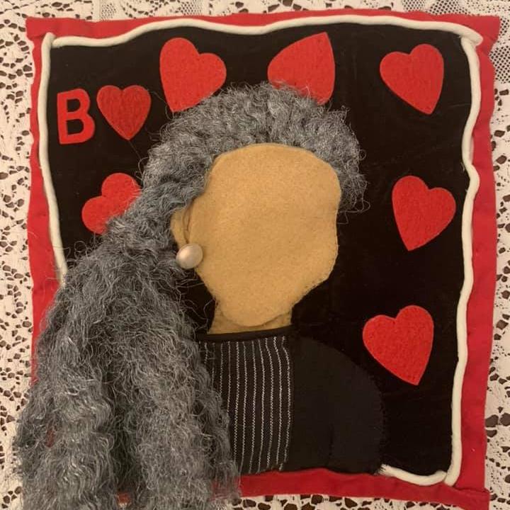 		A red bordered rectangle made of felt with red hearts on it and a blank-faced brown-skinned woman with one pearl earring and long gray hair
	