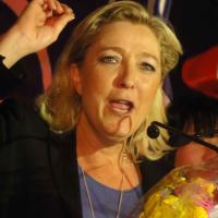 		Marine Le Pen with sholuder-length blonde hair and jacket, with hand upraised in the midst of a speech, with French flag in bakcground
	
