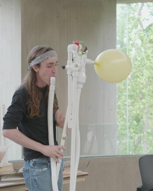 		J Nation blowing on an instrument made out of long white pipes, with a yellow balloon attached
	
