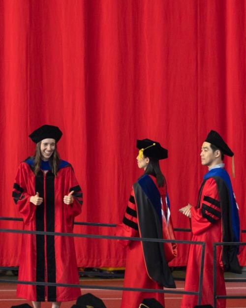 		Several people in long red robes and black caps walk in a line against a red background; one turns to give a thumbs up
	