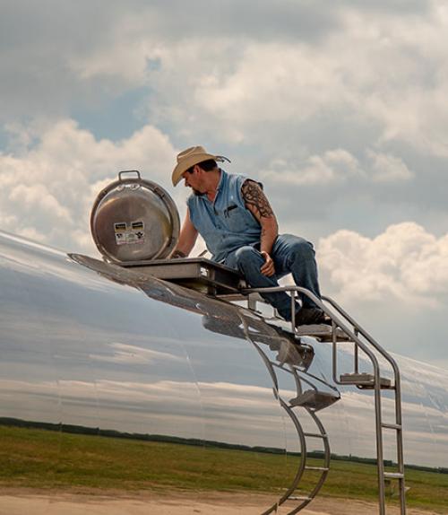 		 Man with a tattoo on top of a tanker truck with the image of the sky reflected off the metal
	