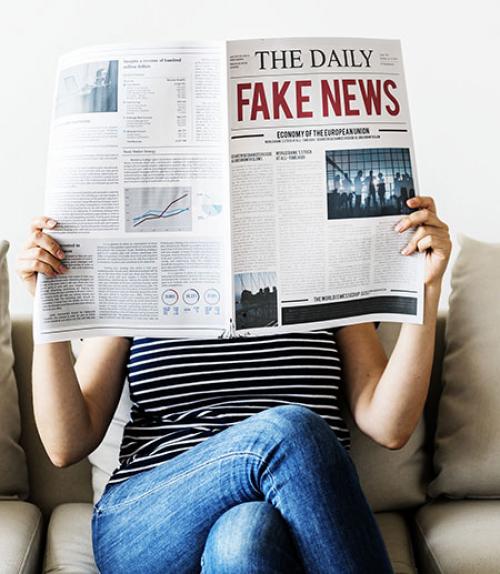 		 woman on couch holding up newspaper with giant headline that says &amp;quot;Fake News&amp;quot;
	