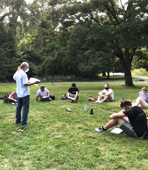 		 A teacher stands in a circle of students sitting on the grass
	