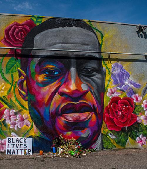 		 Artist drawing of George Floyd&amp;#039;s face on a wall, surrounded by flowers and Black Lives Matter sign
	