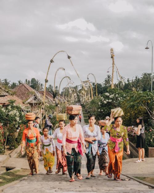Women wearing brightly colored clothes walking in a village with baskets on their heads.