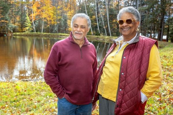 Two people stand in front of a pond surrounded by woods