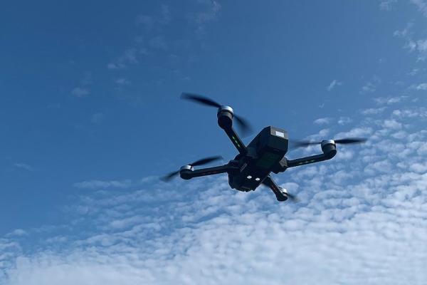 A dark, four-limbed flying drone against a blue sky with fluffy clouds