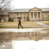 person walks past a puddle that is reflecting a campus building