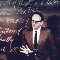 Black and white historical photo of a person in jacket and tie speaking authoritatively in front of a chalk board