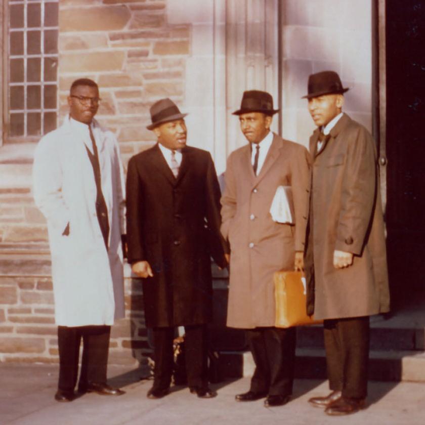 Four people stand in front of a building, wearing dress coats and hats