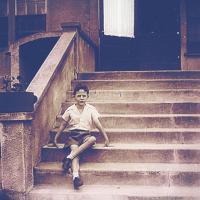 		A boy seated on stone steps, dressed in shorts and a white shirt. Black and white historical image
	