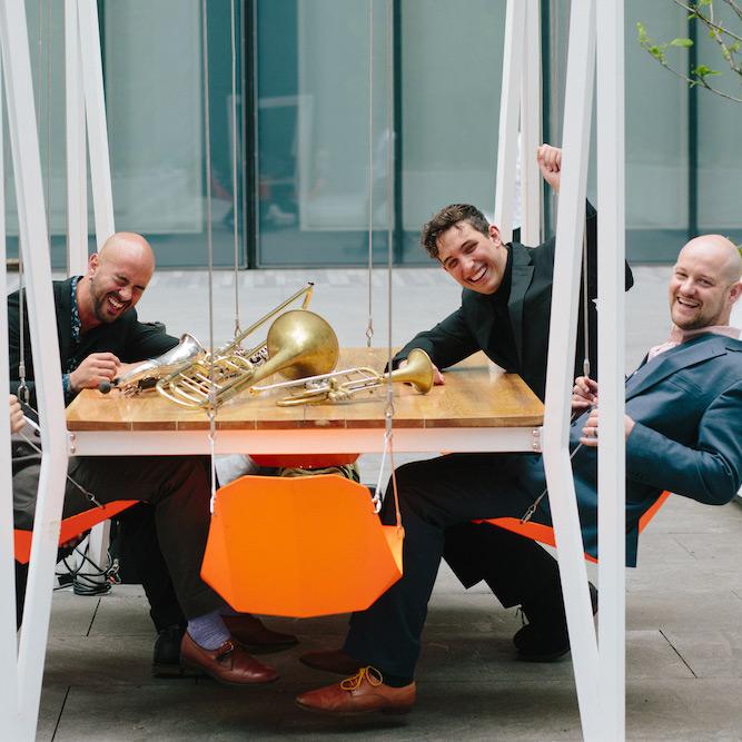 Four people sitting around a table that has musical instruments on it: a saxophone, a trombone and a trumpet