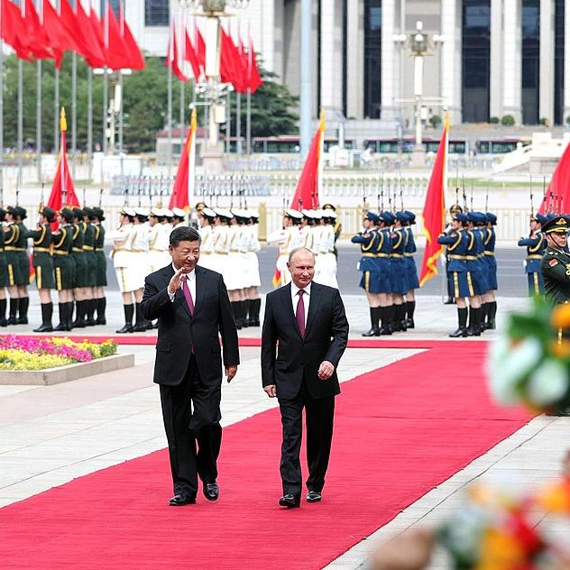 Two people wearing suits walk side by side down a red carpet, waving