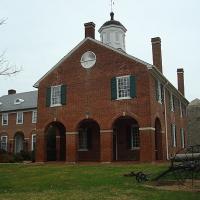 A red brick building with a white painted cupola on top with a weather van, with three large archways in front and a side building. A cannon sits in front.