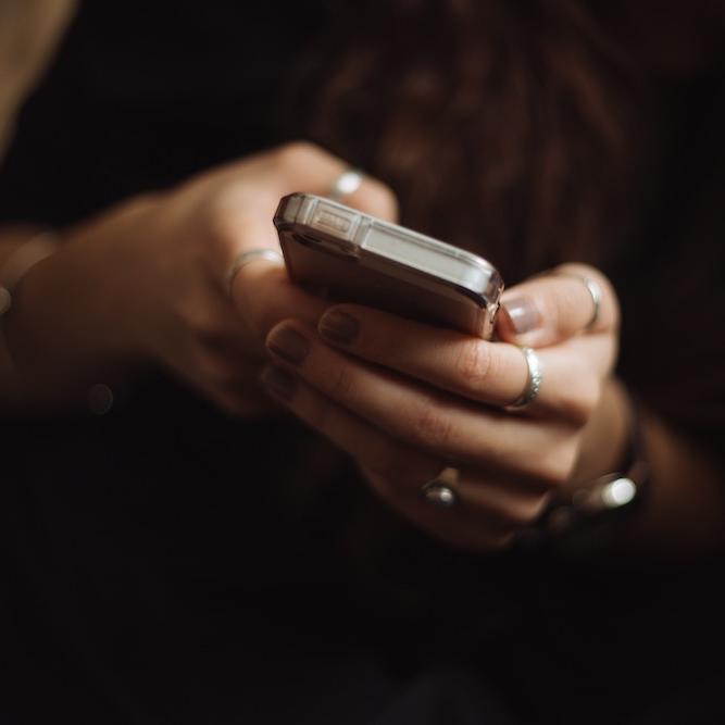 		Two hands (manicured, wearing silver rings) hold a smart phone against a dark backgroun
	