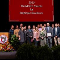 A few dozen people stand on a stage below a banner: 2023 President's Awards for Employee Excellence