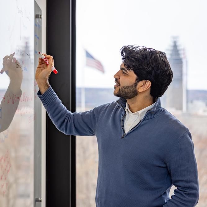 Person writing on a dry-erase board with a window in the background