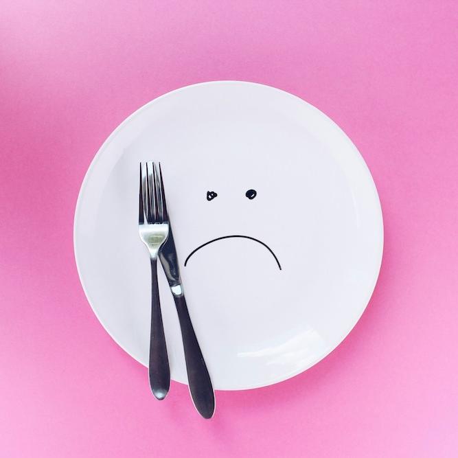 		White plate on a pink background, with a fork and a knife. There is a sad face drawn on the plate
	