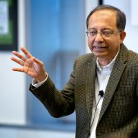 Kaushik Basu wearing a tweed jacket with hand upraised as he delivers a talk.
