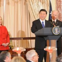 Chinese President Xi Jinping standing at a podium with the US Seal on the front, with Joe Biden behind him and Hilary Clinton to his left dressed in a red pants suit.
