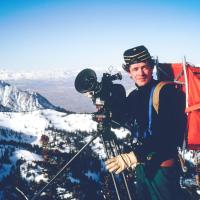 Person in hiking gear with a large camera; mountains in background