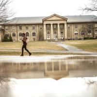 person walks past a puddle that is reflecting a campus building