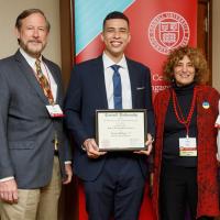 Conor Hodges in suit and tie, smiling and holding his award plaque, flanked by other alumni.