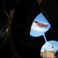 American flag, blue sky and campus building seen through a cutout shaped like an hourglass