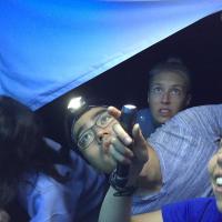 Three people use a flashlight to look at a blue tarp over their heads
