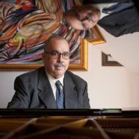 Person wearing a suit, sitting at a piano with a colorful painting in the background