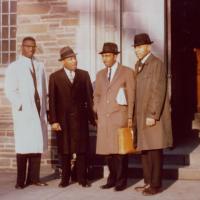 Four people stand in front of a building, wearing dress coats and hats