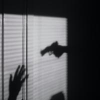 Silhouettes on a wall show a gun aimed at two hands held up in surrender; a scene of nighttime crime