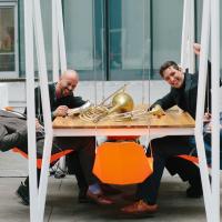 Four people sitting around a table that has musical instruments on it: a saxophone, a trombone and a trumpet