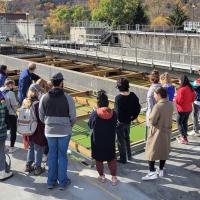 Students standing around an open wastewater treatment facility, which looks like a big cement rectangle with green water and wooden separaters.