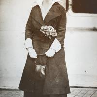 Black and white photo from 1914: a woman in a dark suit and hat highlighted by flowers stands on a wooden dock