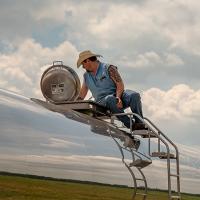 Man with a tattoo on top of a tanker truck with the image of the sky reflected off the metal