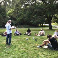 A teacher stands in a circle of students sitting on the grass