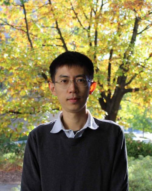 Professor Xin Zhou with sweater on standing in front of a sunshine-lit tree with yellow autumn leaves