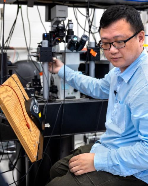Wei Wang, in a blue shirt and black plastic-framed glasses, sits in a lab looking at an instrument while he adjusts another instrument with his right hand.