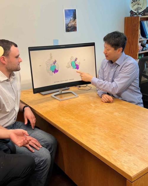 The three researchers are sitting around a desk and Ailong Ke is pointing to an image of the IscB molecule on the computer screen.