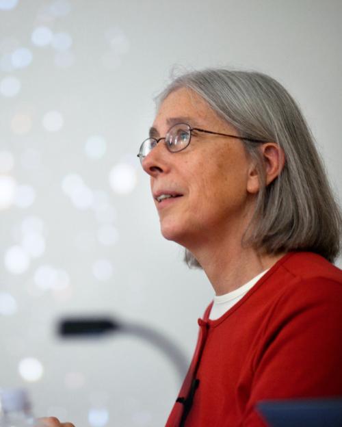Martha Haynes with glasses, shoulder-length gray hair in a red top, with blurred stars on screen behind her