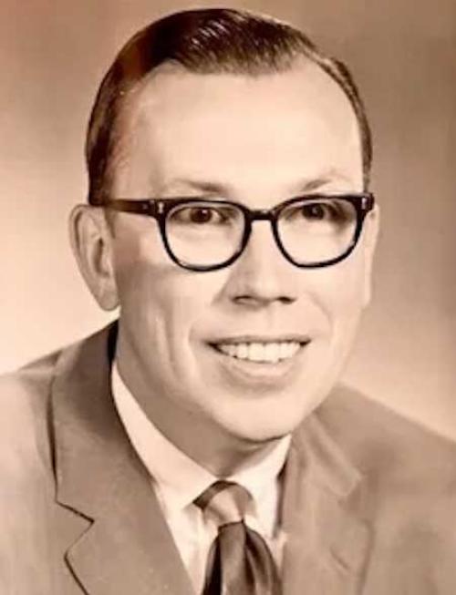 A black and white image of Tom Davis in suit and tie, wearing black plastic glasses and smiling.