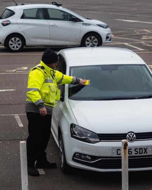 Person wearing a bright yellow jacket places a ticket on a car windshield