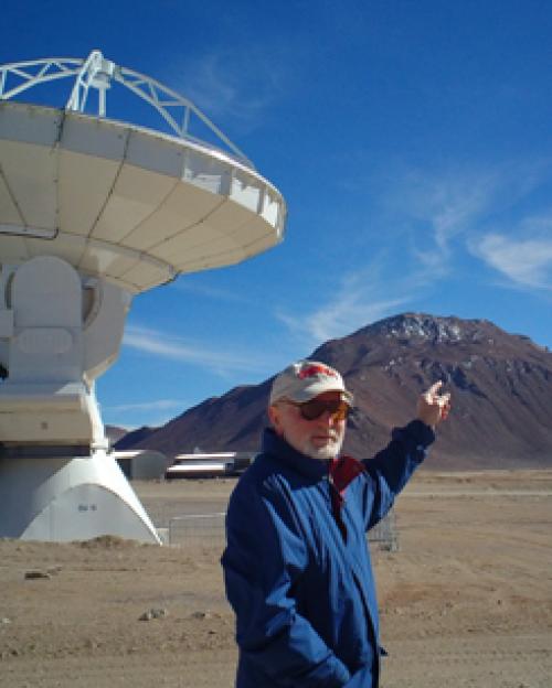 青いジャケット、ベースボール キャップ、サングラス、白ひげのリカルド ジョヴァネッリは、砂漠の山の頂上を指しています。 バック グラウンドで望遠鏡が表示されます。