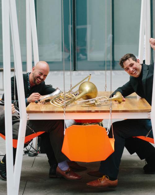 Four people sitting around a table that has musical instruments on it: a saxophone, a trombone and a trumpet