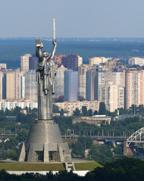 Tall monument in the shape of a figure holding a sword; city buildings in the background