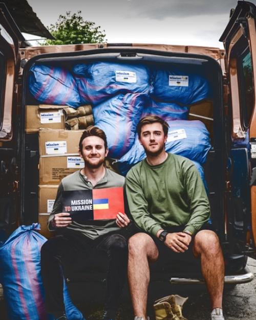 Two people sitting in the back of a van with doors open, showing boxes and bags