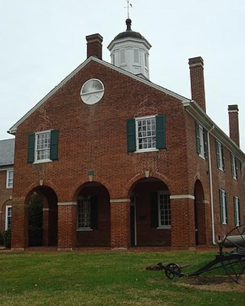 		A red brick building with a white painted cupola on top with a weather van, with three large archways in front and a side building. A cannon sits in front.
	